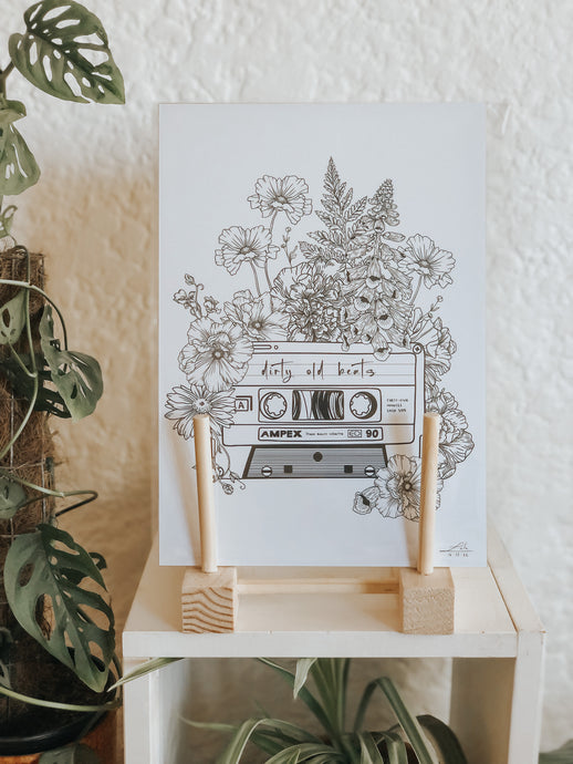 Black and white illustration of a cassette tape surrounded with botanicals. This tape is labelled 
